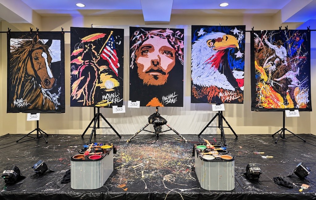 Five Michael Israel paintings on easels: a horse, a cowboy with the American flag, Jesus, an eagle, and a bull rider